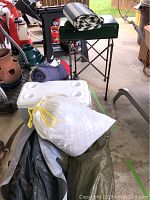 Coleman camp stove on folding stand, cooler, sleeping bags, blankets in bag, and floor mat arranged on floor.