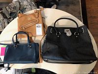Three leather purses on a round white table: large black handbag, medium blue handbag, small tan carry bag.