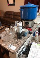 Photo of large blue enamel stockpot on stainless steel electric warmer with glass punch bowl, mugs, water bottle, and assorted kitchen items on a granite countertop with brown couch and map visible in background.