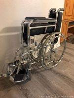 Folded 24 inch foldable wheelchair with silver metal frame, black seat and backrest, large rear wheels and small front caster wheels.