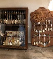 Souvenir spoons in two wooden display cases, one with a glass door, showing souvenir spoons of varied styles and motifs.