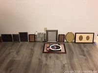12 picture frames of various sizes and finishes arranged on a wood floor against a wall. Several frames lean against the wall, others stacked flat. Frames include wood tones, silver, gold, and white colors. One has a double oval mat.