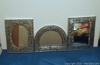 Three assorted Mexican tin pressed mirrors displayed side by side on a flat surface. The left and right mirrors are rectangular with ornate punched tin frames, the center mirror is arch-shaped with matching decorative punched tin edges.
