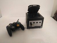 Front view of black Nintendo GameCube console with open disc tray and connected wired controller
