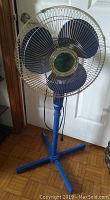 Front view of blue floor fan showing metal wire cage, three blades, and bent foot on base