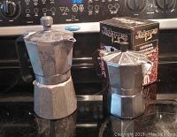 Closed view of large and small aluminum stove top coffee makers on black stove surface, small is boxed