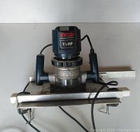 Front view of Ryobi router with 1.5 HP marking, dual handles, and two horizontal metal bars clamped to a flat base