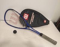 Blue Wilson squash racket with grey grip handle, black squash ball and black Wilson branded cover on white background.