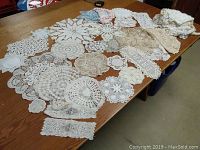 Over 50 assorted small, medium, and large crochet and lace doilies spread out on a wooden table showing various patterns and sizes.