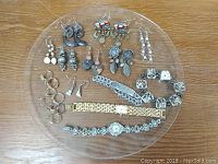 Clear round tray shows full assortment of watches, earrings, rings, and bracelet laid out for display on wood surface