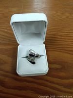 Ring shown inside white jewelry box on wooden surface, visible inlay and stones.