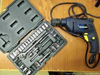 Blue and black Mastercraft electric drill with cord lying next to a black plastic case containing 3/8" drive socket set with various sockets and accessories, some sockets missing.