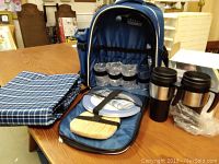 Picnic backpack with removable wine cooler holding four glasses, two stainless steel travel mugs, white plates, wooden cutting board, and blue checkered tablecloth.