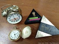 View of gold colored Bulova alarm clock, ornate pocket watch, Olde Tower sealing wax box with metal peace symbol stamp and three wax sticks
