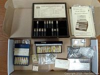 Photo showing cardboard box containing vials labeled Gruen Watch Makers Guild with multiple small small vials and boxes of watch screws and parts in plastic bags.