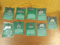 Nine pewter building collectibles arranged in rows on wood surface, each with a green informational card with white text and red ribbon on top