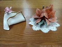 Two Capodimonte porcelain flower pieces on a wooden surface with a white lace doily underneath the larger piece.