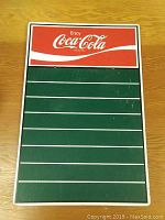Front view of a rectangular Coca Cola chalkboard sign with red header and green writing area with white lines.