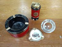 Photo of four items: a large red and black ashtray, a red and black Ronson lighter, a clear round glass ashtray, and a small ceramic ashtray with painted design.