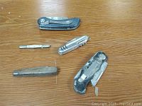 Five various pocket knives laid out on a wooden surface, showing different designs and degrees of wear.