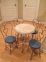 Full view of child's bistro set with round marble top table and four wrought iron blue chairs with heart backs on wood floor.