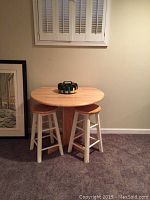 Photo shows a wooden pedestal dinette table with two sides that can be lowered or raised, along with two matching wooden stools with white legs and natural wood seats.