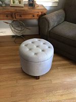 Grey round fabric ottoman with button tufted removable lid, hardwood floor background, next to a wooden desk and grey fabric armchair.
