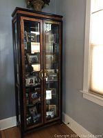 Front view of tall Pulaski wall unit with burled wood, ebony trim, glass front double doors revealing multiple framed photographs on glass shelves inside, gold-toned handles, standing against a gray wall near a window.