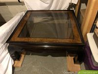Image of a large square coffee table with a dark ebony base and burled wood frame surrounding a glass inset top. The table is positioned indoors on a concrete floor.