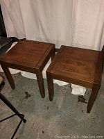 Pair of wooden square end tables shown from angled side view on concrete floor with white backdrop.