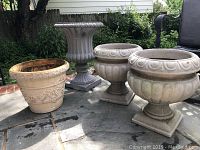 Four assorted lightweight resin outdoor planting pots placed on stone patio surface, with greenery and part of house and chair visible in background.