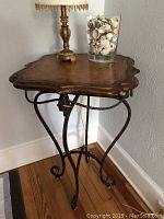 Full view of the decorative table showing the wooden ornate top and scrolled iron legs with a lamp and decorative glass vase on top.