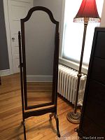 Full length standing mirror with dark wooden frame, decorative top edge, and pivoting stand on legs, placed indoors near lamp and radiator.