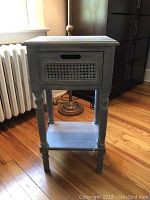 Front view of grey wooden side table with single drawer featuring cane panel and lower shelf, placed on wooden floor near radiator and lamp.