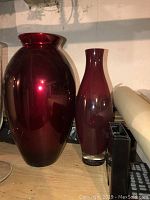 Two red glass vases side by side against a white wall, the larger on the left measuring 14 1/2 inches, the smaller on the right 12 1/2 inches.