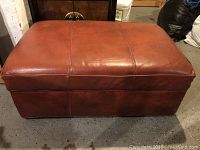 Front view of rectangular brown leather ottoman showing some wear on corners and stitch detailing.