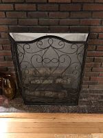 Black metal folding fireplace screen with ornate scrollwork and mesh backing in front of a brick fireplace.