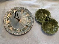 Photo showing the top view of both dishes, one large round with floral and heart design and metal handle, one smaller double shell-shaped green glazed dish with handle