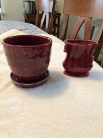 Two red pottery vases on a table - a larger round vase with floral texture and saucer, and a smaller uniquely shaped vase