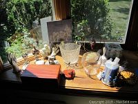View of the collection of Snowbabies figurines, Lane cedar box, various trinket boxes, candy dish, bell, and candle displayed on a wooden windowsill with garden view.