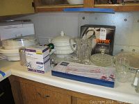 Overview of kitchen countertop with all lot items arranged including bread maker, Pyrex box, glassware, tureen, and packaged item.
