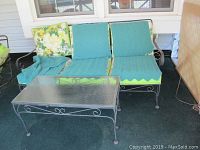 Front view of metal framed patio sofa with green cushions, floral pillow, and two blankets, alongside metal framed glass top patio table