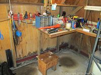 Workbench with various hand tools, hardware items, boxes, and small stool in a workshop space