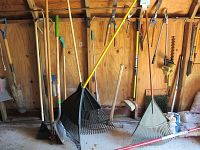 Photo showing several long-handled yard tools hanging on wall including different types of rakes, shovels, and pruning tools