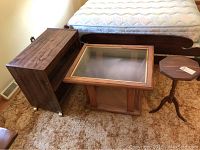 Three wooden furniture pieces arranged indoors on carpet. Includes a wood rolling stand with caster wheels on left, glass top side table in center, and small wooden plant stand on right.