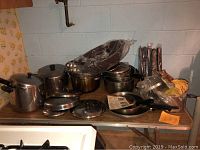 Photo showing assortment of stainless steel pots, pans, some with lids, neatly stacked with several utensils still in plastic bags on folding table