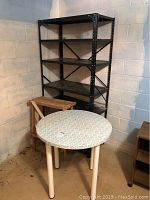 Full view showing black metal shelf with four shelves, round table with patterned contact paper top, and wooden slatted shelf