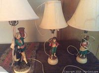 Three ceramic figurine base table lamps with beige fabric shades on a wooden surface.