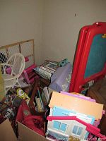 Photo showing corner with white wicker chair, toy dollhouse, art easel, and stacks of books and games including Risk board game.