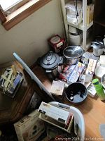 Wide shot of floor and shelf showing multiple kitchen appliances including pressure cooker, crock pot, scale, bakeware boxes, and plastic storage bins.